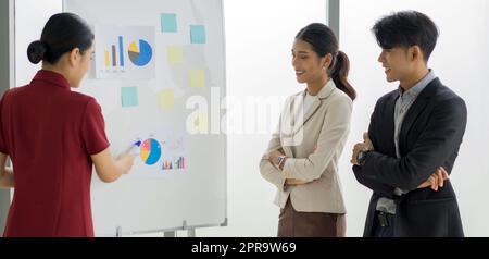 Junge asiatische Geschäftsfrau in braunem Anzug mit Blick auf das Tortendiagramm auf der weißen Tafel. Auf dem Präsentationboard befinden sich Notizen und Papiere. Stockfoto