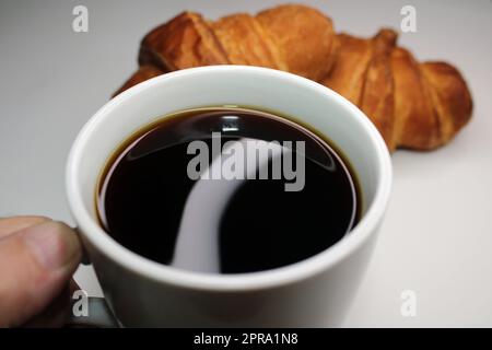 Weiße Tasse mit Nahaufnahme des schwarzen Kaffees auf hellem Hintergrund. Im Hintergrund gibt es zwei Croissants. Kochen, Selbstbacken Stockfoto