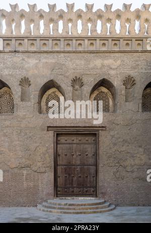 Verwitterte Holztür, perforiertes Stuckfenster mit Blumenmustern und drei Stufen an Steinmauern Stockfoto