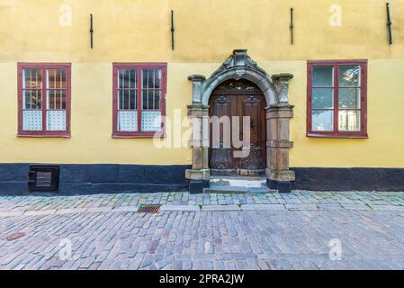 Dekorierte Holztür mit Steinrahmen, schmiedeeisernen Holzfenstern in gelber Wand und Kopfsteinboden Stockfoto