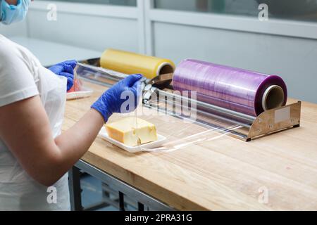 Die Frau wickelt ein Stück Käse, das auf einer Plastikschale liegt, in einen durchsichtigen Lebensmittelfilm. Stockfoto