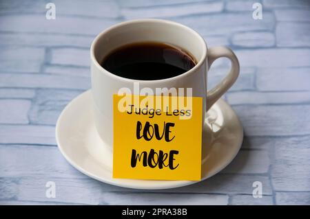 Inspirierender Zitattext auf gelbem Notizblock auf der Kaffeetasse – Judge less love more. Motivationskonzept Stockfoto