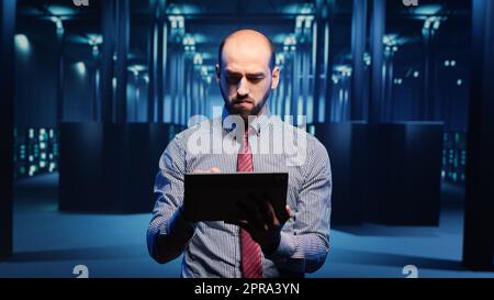 Männlicher Infrastrukturspezialist, der ein Tablet für die Hardware-Digitalisierung einsetzt und Cloud-Computing und Cluster-Computer untersucht. Rechenzentrumstechniker, der Serverdiagnosen durchführt. Stativaufnahme. Stockfoto