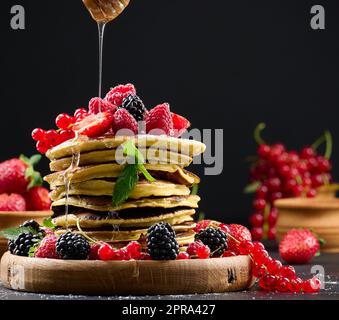 Stapel von Pfannkuchen mit frischem Obst bestreut mit Honig auf schwarzem Hintergrund Stockfoto