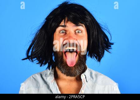 Ein komischer bärtiger Mann mit schwarzer Perücke zeigt Zunge. Glücklicher Mann mit Bart und Schnurrbart in Perücke. Menschliche Emotionen. Gesichtsausdruck. Gefühle und Emotionen. Hipster Stockfoto