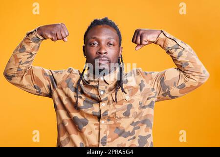 Sportlicher junger Erwachsener, der vor dem Training im Studio Bizepskraft auf gelbem Hintergrund zeigt. Ein afroamerikanischer Sportler, der Arme zeigt, Muskeln aus Fitness Stockfoto