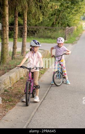 Zwei Mädchen im Grundschulalter, Kinder, die Fahrrad fahren, Fahrrad fahren, auf einer Landstraße in einer ländlichen Gegend zusammen Freunde, Schwestern, Geschwister sp Stockfoto