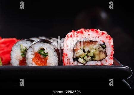 Japanische Reisbrötchen Uramaki und Maki mit Lachs, Gurke und rotem Kaviar auf einem schwarzen Teller. Stockfoto