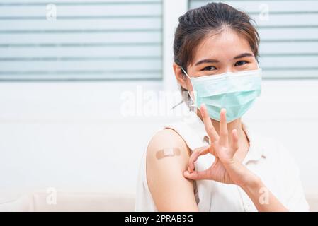 Frau lächelt, sie zeigt auf Pflaster am Arm, den sie geimpft hat, und zeigt OK Finger Stockfoto