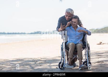 Ältere Frau im Rollstuhl sitzt und Ehemann ist ein Rollstuhl Benutzer Smartphone macht Selfie am Strand Stockfoto