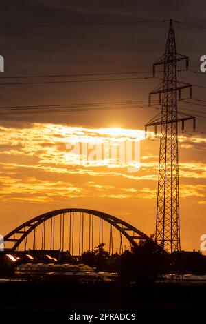 Der goldene Himmel mit Sonnenstrahlen und Linsenerscheinungen zeigt Solarenergie mit der Pylonsilhouette des Stromturms in goldenem Sonnenuntergang und orangefarbenem Himmel für nachhaltige Energie oder erneuerbare Ressourcen von der Dämmerung bis zur Dämmerung Stockfoto