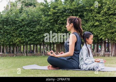 Mutter übt Yoga-Übungen mit ihrer Tochter im Freien in meditierender Pose zusammen Stockfoto