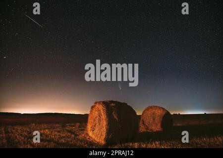 Meteor und Comet Neowise C2020 F3 in Night Starry Sky über Heuhaufen auf dem landwirtschaftlichen Sommerfeld. Nachtstars Über Der Ländlichen Landschaft Mit Heuballen Nach Der Ernte Stockfoto