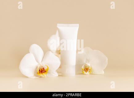 Weiße cremefarbene Röhre in der Nähe von weißen Orchideenblumen auf hellbeige Nahaufnahme. Modell. Hautpflegeprodukt Stockfoto