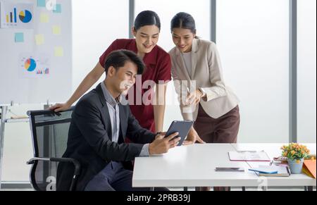 Junger asiatischer Geschäftsmann im Anzug mit Tablet-PC, während beide Kollegen an den Informationen auf dem Bildschirm interessiert waren. Diagramm, Diagramm und Notiz befinden sich auf dem Präsentationstafel im Hintergrund. Stockfoto