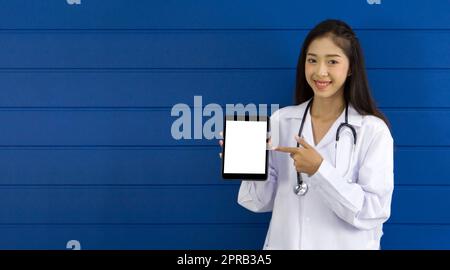Junger asiatischer Arzt in weißem Kleid und Stethoskop, der mit dem Finger auf einen Tablet-Computer mit weißem Bildschirm zeigt, der vor einer blauen Wand steht. Stockfoto