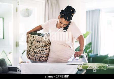 So viel zu tun und so wenig Zeit. Eine junge Frau, die zu Hause am Telefon Kleidung bügelt. Stockfoto