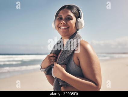 Übung bringt das Beste in mir heraus. Eine sportliche junge Frau, die am Strand Kopfhörer und ein Handtuch um den Hals trägt. Stockfoto