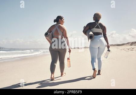 Wir haben es uns zur Priorität gemacht, regelmäßig Yoga zu praktizieren. Zwei junge Frauen, die mit ihren Yogamatten am Strand spazieren. Stockfoto
