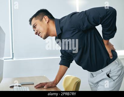 Warum muss mein Rücken zuerst altern. Ein hübscher junger Geschäftsmann, der allein in seinem Büro steht und unter Rückenschmerzen leidet. Stockfoto