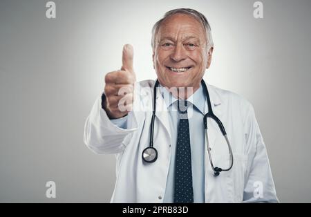 Jahre später liebe ich immer noch meinen Job. Ein älterer, männlicher Arzt in einem Studio, der vor grauem Hintergrund Daumen nach oben gibt. Stockfoto