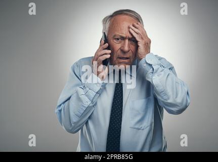 Oh mein... was machen wir jetzt? Ein älterer Geschäftsmann, der allein vor einem grauen Studiohintergrund steht und gestresst aussieht, während er sein Handy benutzt. Stockfoto