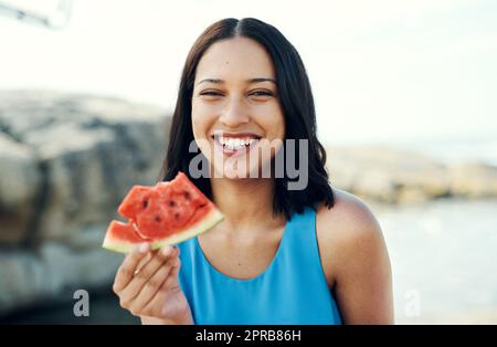 Hmm nichts schlägt frische Wassermelone. Eine junge Frau, die am Strand ein Stück Wassermelone isst. Stockfoto