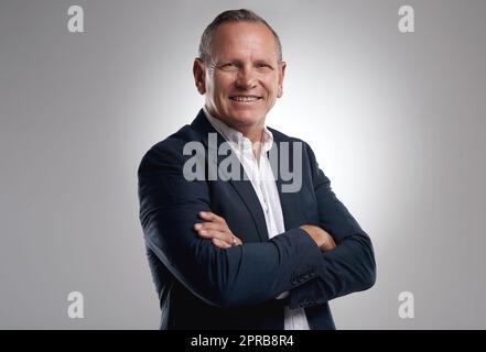 Ein Spielwechsel in einer Welt voller Spieler. Ein gutaussehender, reifer Geschäftsmann, der allein vor grauem Hintergrund im Studio mit gefalteten Armen steht. Stockfoto