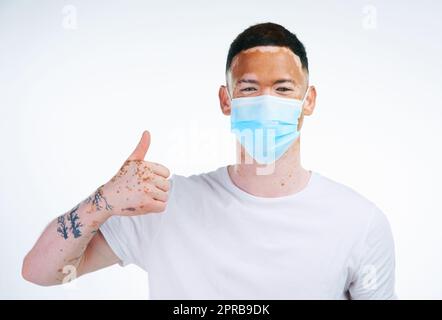 Indem ich mich um mich selbst kümmere, kümmere ich mich um Du auch. Portraitaufnahme eines jungen Mannes mit Vitiligo, der eine schützende Gesichtsmask trägt und auf weißem Hintergrund posiert. Stockfoto