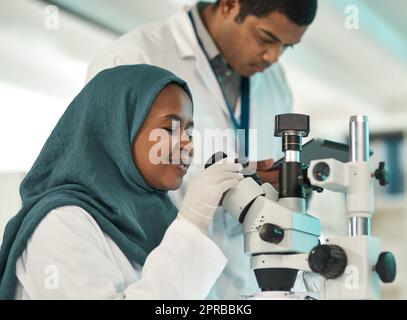 Durchführung kontrollierter Tests. Ein junger Wissenschaftler, der ein Mikroskop benutzt, während er mit einem Kollegen in einem Labor zusammenarbeitet. Stockfoto