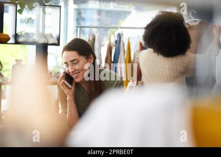 Sie sind jederzeit willkommen. Eine Frau, die auf ihrem Handy spricht, während sie in einem Bekleidungsgeschäft arbeitet. Stockfoto