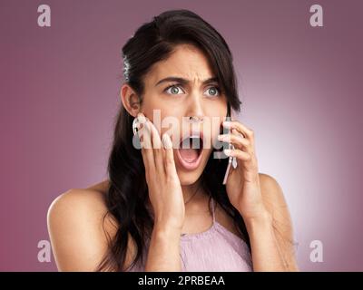 Freundschaft ist Tratschen wie Schulmädchen. Eine schöne junge Frau, die schockiert aussah, mit der Hand an der Wange, während sie vor einem rosa Hintergrund mit einem Mobiltelefon sprach. Stockfoto