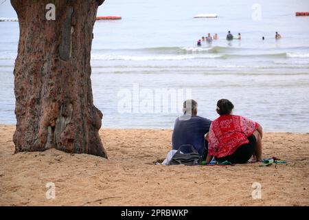 Ein älterer Mann und eine ältere Frau sitzen zusammen am Strand, neben einem Baum, und schauen auf das Meer in Jomtien, Pattaya, Thailand. Entspannen, entspannen, Urlaub, Urlaub Stockfoto