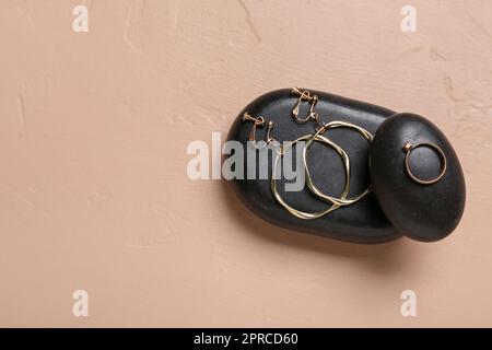 Schwarze Steine mit goldenen Ohrringen und Ring auf beigefarbenem Hintergrund Stockfoto