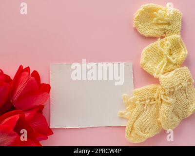 Rote Tulpenblüten, gelbe Stiefel und Handschuhe für ein Neugeborenes auf weißem Hintergrund mit Kopierbereich. Muttertag Grußkarte. Wünsche für Mutter schreiben. Stockfoto