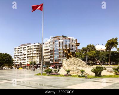 Platz im Stadtzentrum mit weißen Häusern. In der Nähe des Zentrums befindet sich eine rote Flagge der Türkei auf einem hohen Fahnenmast. Auf dem Platz befindet sich ein Bronze-Denkmal. Stockfoto