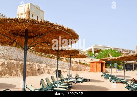 Tropischer Sandstrand im Urlaub, ein tropisches Resort mit Sonnenliegen, Sonnenliegen und Sonnenschirmen in Form von Strohhüten im Hintergrund der s Stockfoto