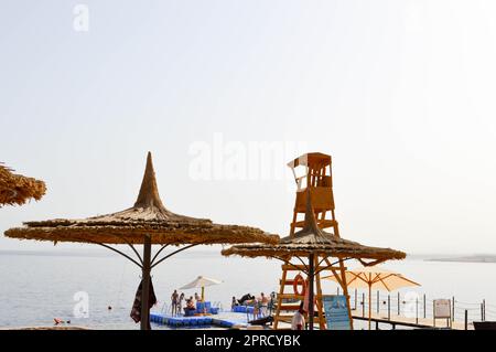 Wunderschöne Strohschirme in Form von Hüten und grünen Palmen in einem tropischen Wüstenresort vor einem blauen Salzmeer, schwimmende Hosen Stockfoto