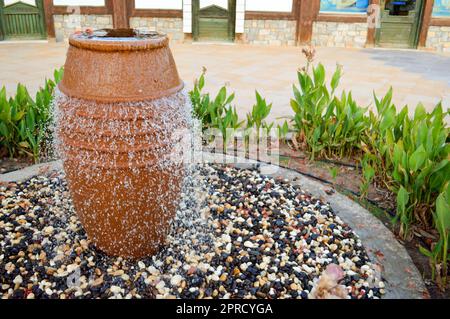 Ein schöner kleiner Brunnen in Form einer braunen Vase, ein Krug mit herabfallenden Wassertropfen auf farbigen Steinen, der in einem Blumenbeet mit grüner Pflanze steht Stockfoto