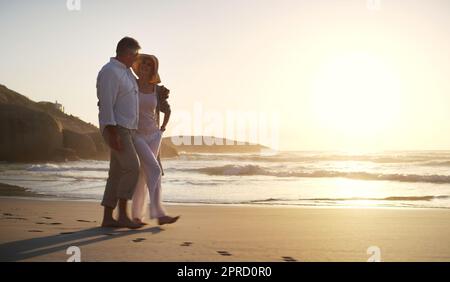 Die Sonnenuntergänge erinnern mich an die Zeit, als wir uns das erste Mal trafen. Die ganze Länge des Bildes eines liebevollen Seniorenpaares, das bei Sonnenuntergang am Strand entlang spaziert. Stockfoto