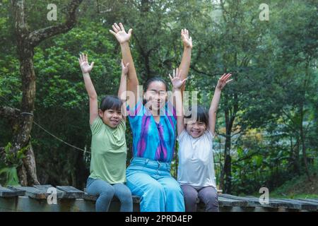 Glückliche junge Mutter mit kleinen Mädchen, die im Sommer auf einer Holzbrücke in der Nähe des Flusses sitzen. Mutter und zwei Töchter haben Spaß, während sie Zeit miteinander verbringen Stockfoto