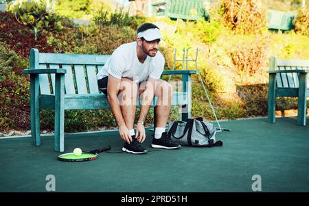 Auf dem Platz steht ein sportlicher junger Mann, der seine Schnürsenkel schnürt, während er auf einer Bank auf einem Tennisplatz sitzt. Stockfoto