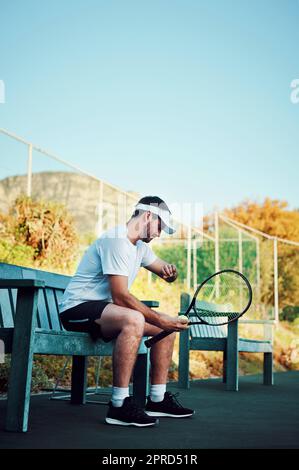 Das Spiel beginnt bald. Ein sportlicher junger Mann schaut auf seine Uhr, während er auf einer Bank auf einem Tennisplatz sitzt. Stockfoto