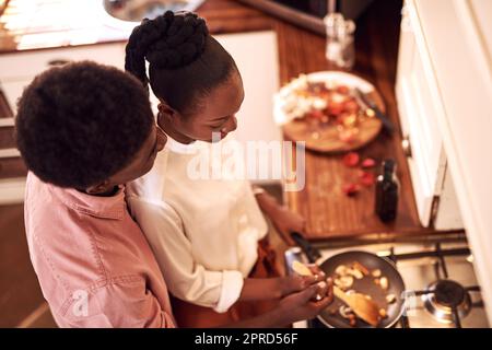 Braucht Hilfe, Baby. In einem Hochwinkel wird ein anhänglicher junger Mann gedreht, der seine Frau umarmt, während er zu Hause in der Küche kocht. Stockfoto