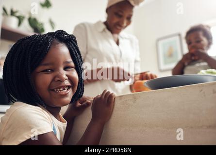 Meine Mutter macht die besten Mahlzeiten aller Zeiten. Porträt eines entzückenden kleinen Mädchens, das in der Küche steht, während ihre Mutter und ihre kleine Schwester im Hintergrund stehen. Stockfoto