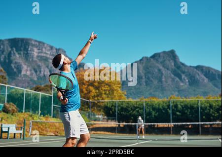 Timing ist alles in diesem Sport. Ein junger männlicher Tennisspieler, der sich bereit macht, den Ball auf einem Tennisplatz im Freien zu servieren. Stockfoto