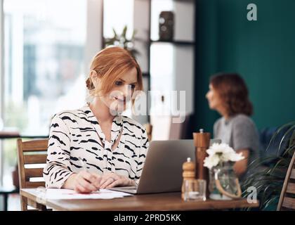 Ein paar wichtige Dinge muss ich aufschreiben. Eine attraktive Frau mittleren Alters, die tagsüber an ihrem Laptop arbeitet und sich in einem Café Notizen macht. Stockfoto