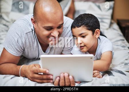 Ich Frage mich, was als Nächstes passiert. Ein fröhlicher junger Mann und sein Sohn schauen sich gemeinsam Videos auf einem digitalen Tablet an, während sie tagsüber auf einem Bett zu Hause abhängen. Stockfoto