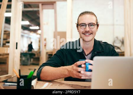 Eine warme Tasse Kaffee bringt mich in den Tag. Porträt eines hübschen jungen Zimmermanns, der Kaffee trinkt und in seiner Werkstatt an einem Laptop arbeitet. Stockfoto