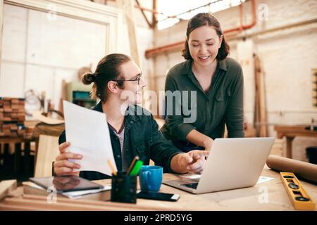 Die Planung ist in jeder Phase des Erfolgs von grundlegender Bedeutung. Zwei junge Tischler planen und arbeiten gemeinsam an einem Laptop in ihrer Werkstatt. Stockfoto